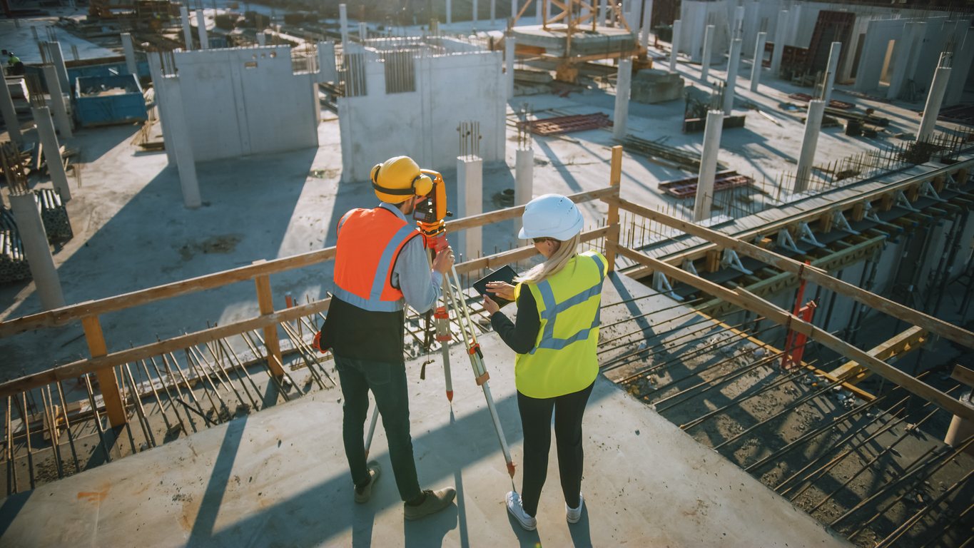 Construction Worker Using Theodolite Surveying Optical Instrument for Measuring Angles in Horizontal and Vertical Planes on Construction Site. Engineer and Architect Using Tablet Next to Surveyor.
